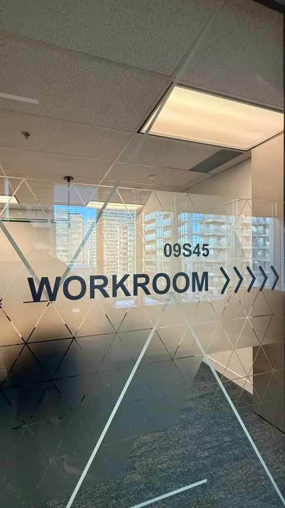 A glass office door featuring a frosted vinyl privacy film with a geometric triangle pattern and custom black lettering that reads 'WORKROOM 09S45' with directional arrows.