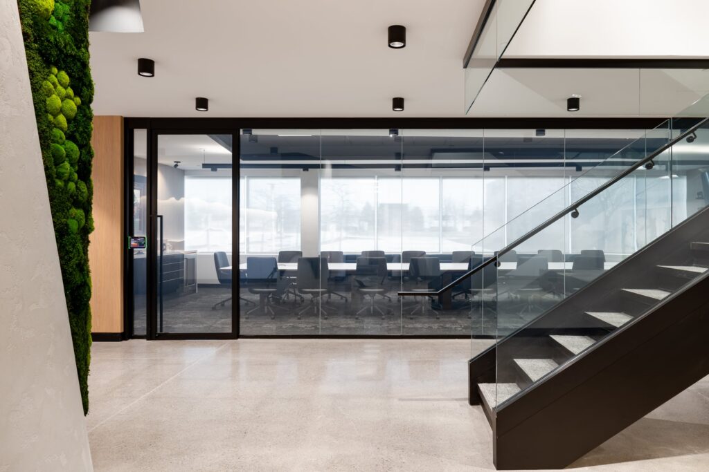 View through a glass conference room partition toward a modern staircase and a vibrant green moss wall accent.