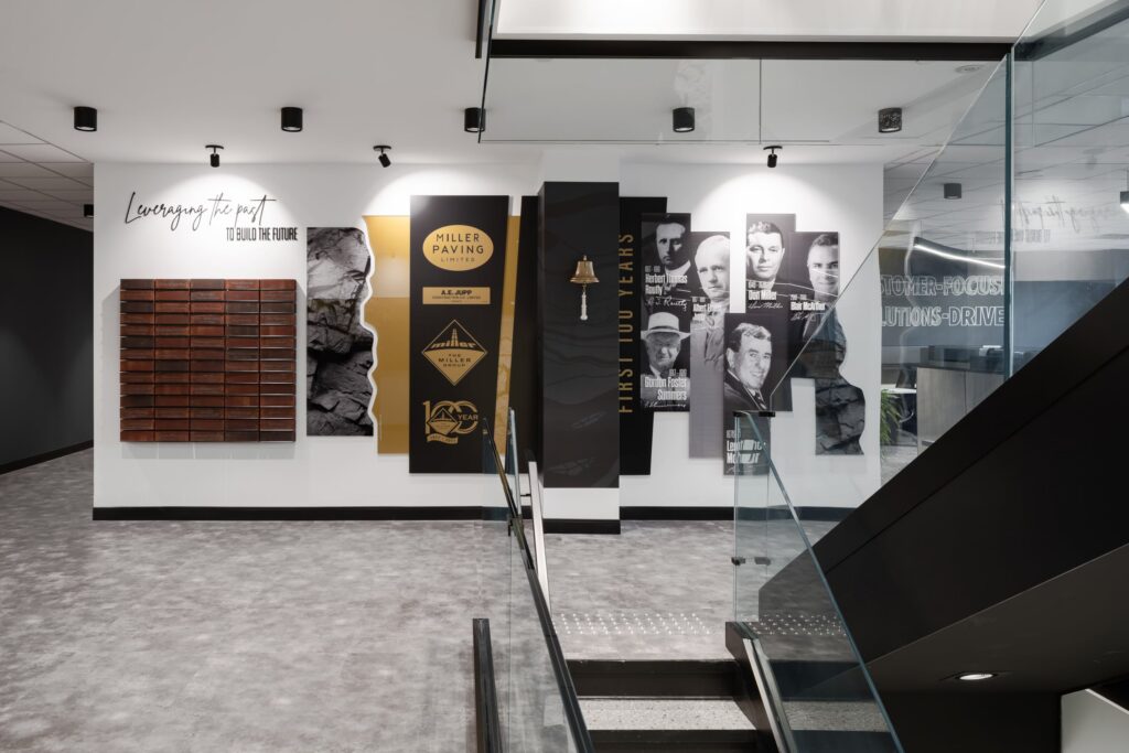 A multi-layered corporate history wall titled "Leveraging the Past to Build the Future" featuring dimensional portraits, a physical bronze bell, and etched brand milestones.