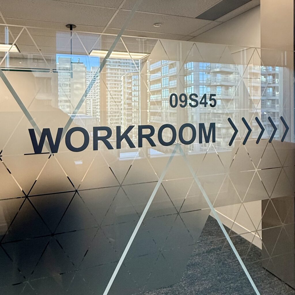 A glass office partition featuring a custom frosted window film with a geometric triangle pattern and black directional wayfinding graphics for a workroom.